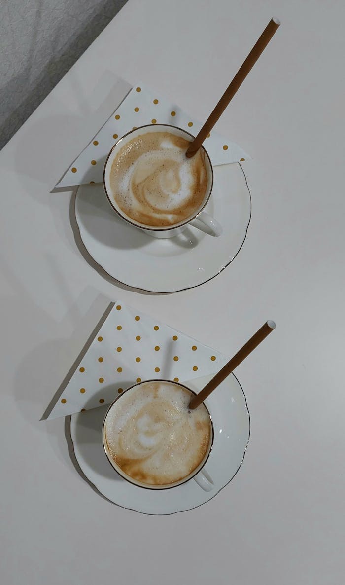 Two cappuccino cups with straws on a white table, featuring polka dot napkins.