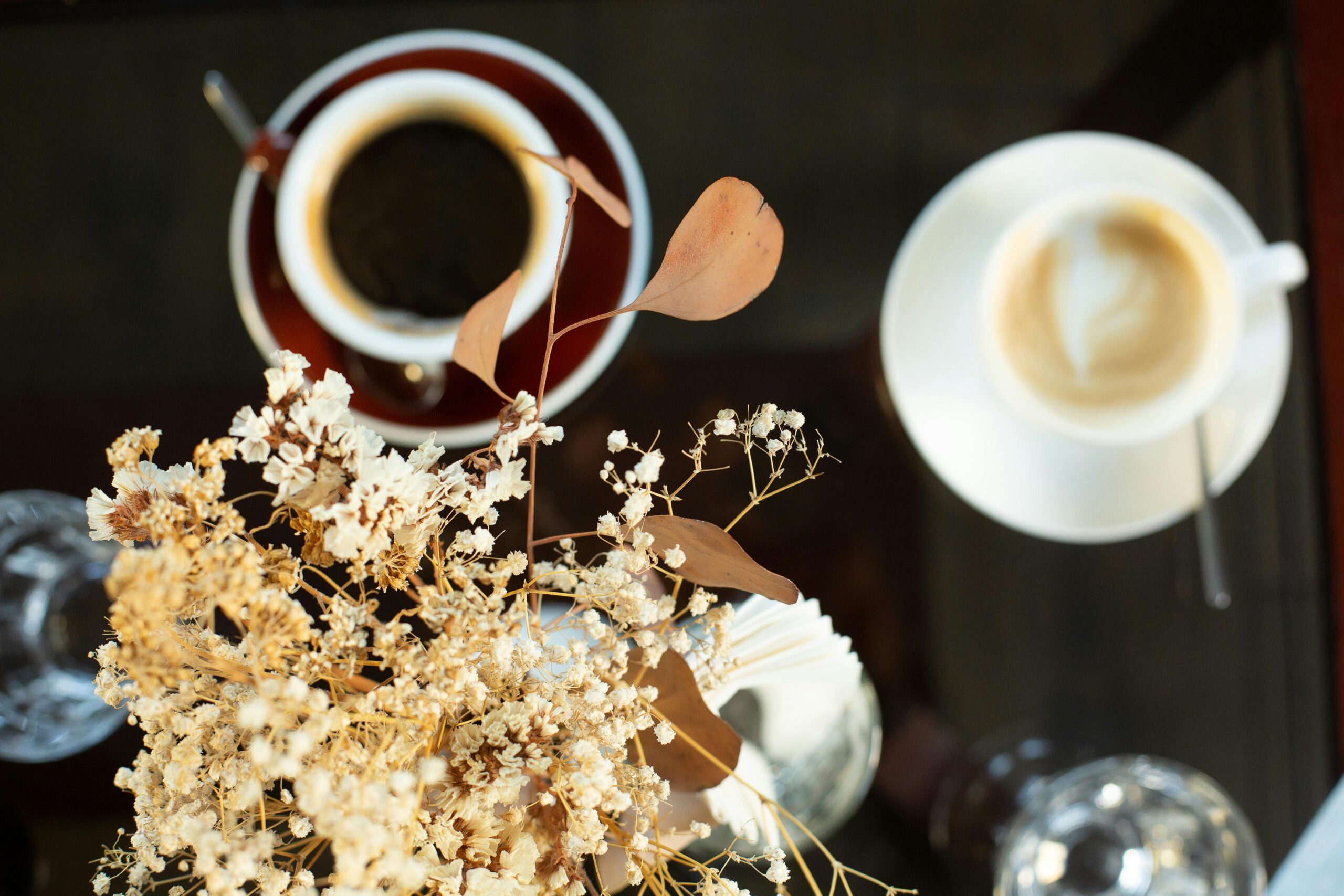 Top view of black and latte coffee with dry floral decor and vase, creating a serene cafe vibe.